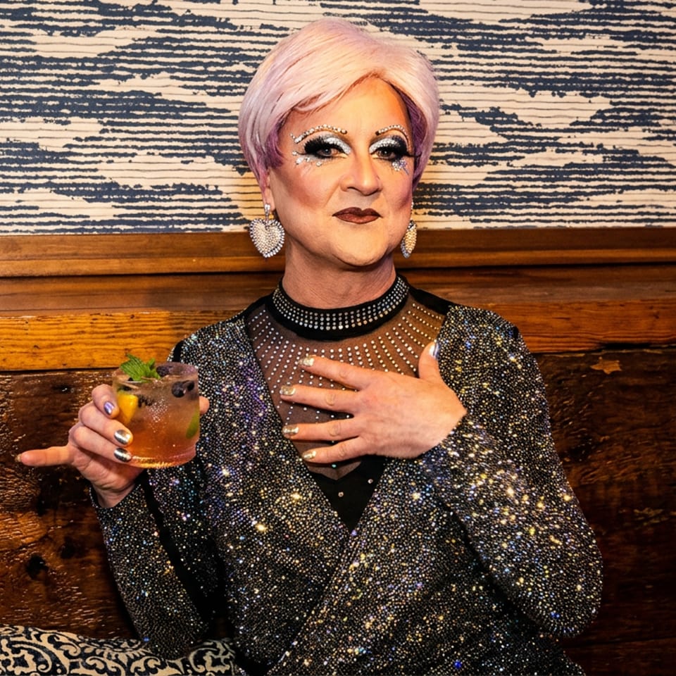 Drag performer in a sparkly outfit and pink wig, holding a cocktail in a stylish bar.